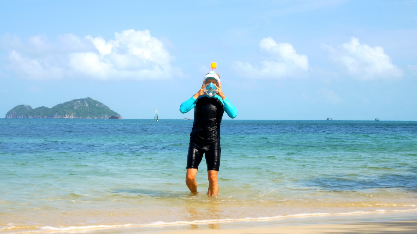 Man in full-face snorkeling mask steps out of tropical sea onto sandy beach