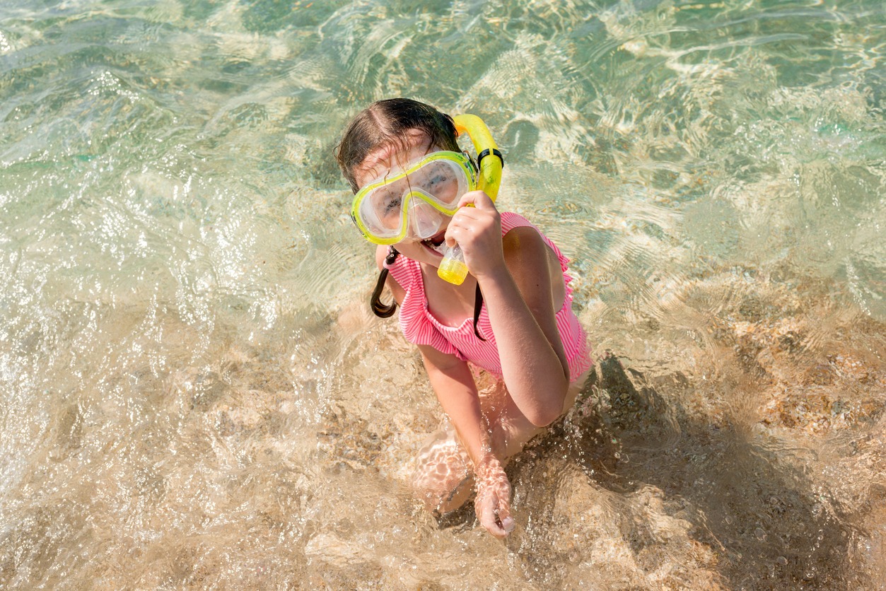 Top view of girl in pink swimsuit with snorkeling mask sitting on sandy beach