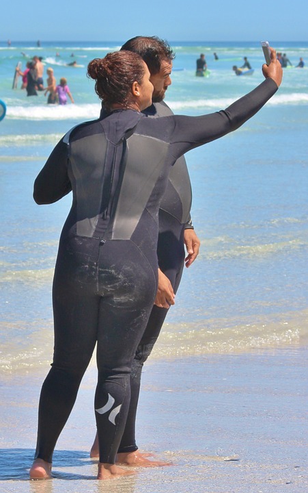 a man and a woman wearing wetsuits taking a photo at the beach