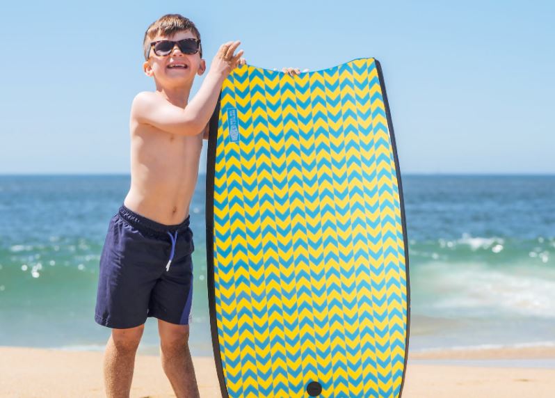 a boy wearing blue shorts and sunglasses holding a skimboard on the beach