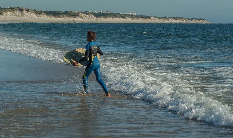 a boy with his back turned holding a skimboard at the shoreline