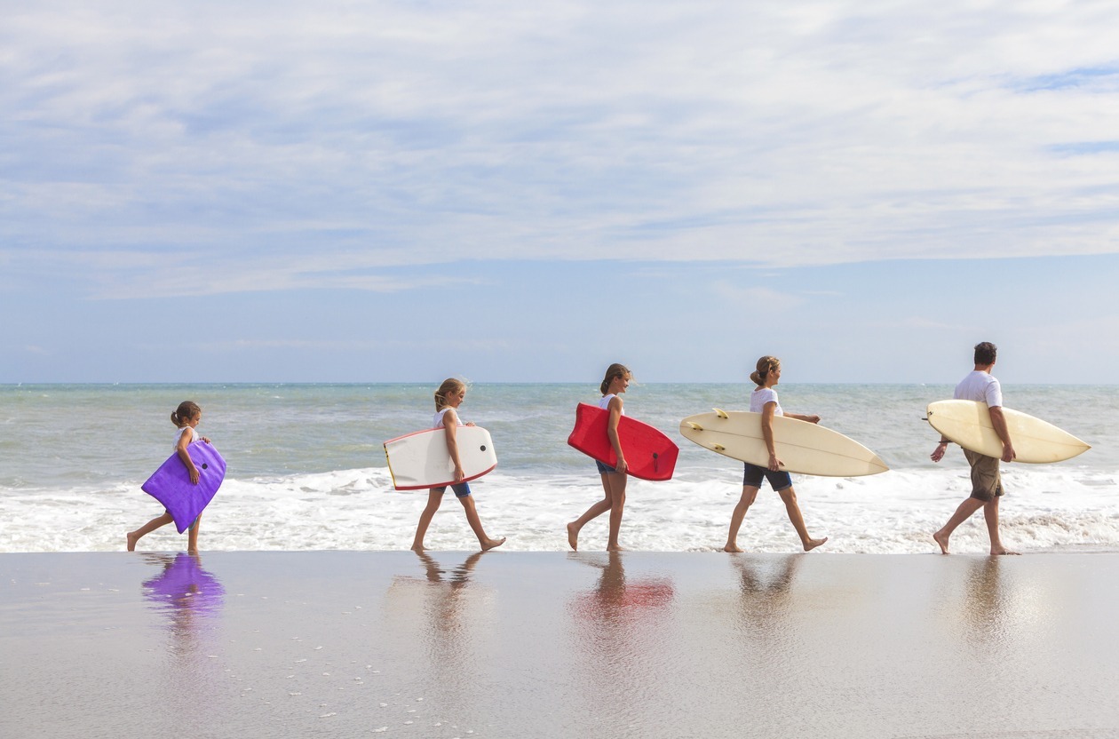 the rear view of a family- mother, father, and female children going surfing with surfboards and bodyboards