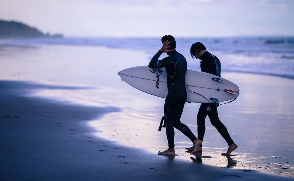 two men walking out of the sea wearing wetsuits and carrying a surf board